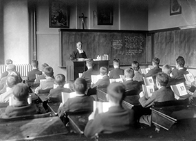 An older Christian Brother, in his collar and robe, sits at an elevated desk looking out at his classroom of young boys, all dressed neatly in jackets and seated at their desks reading the same page in the same books.  We see the back of the boy’s heads.